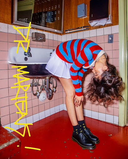 Young woman in a red and blue striped shirt and white skirt is leaning over in a bathroom with pink tile and a white sink.