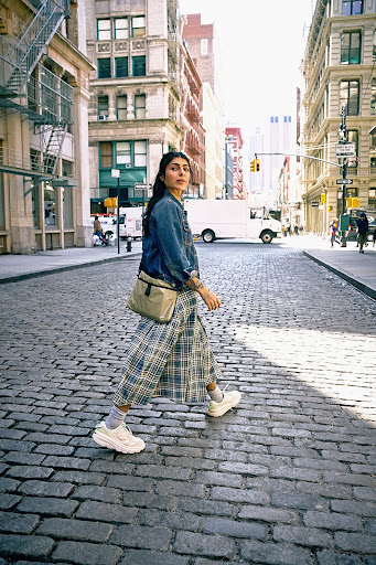 A woman walks across a city street in a denim jacket, plaid skirt, and sneakers, with buildings and a white van in the background.