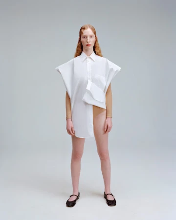Model with red hair standing in an oversized white shirt and dark flat shoes against a plain grey background