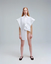 Model with red hair standing in an oversized white shirt and dark flat shoes against a plain grey background