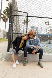 Young couple sitting against a chain-link fence outdoors, styled by Heather Rest with casual denim and outerwear.