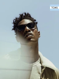 Low-angle portrait of a Black man wearing dark sunglasses and a tan jacket against a clear blue sky background.