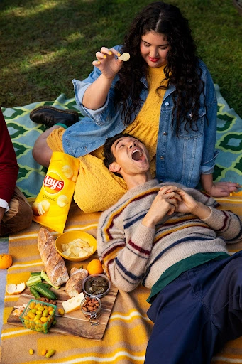 Two people having a picnic on a blanket with a bag of Lay's potato chips and various snack foods laid out.