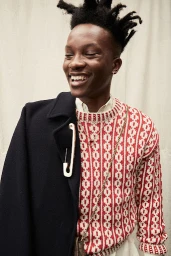 Portrait of a smiling young Black man wearing a red patterned knit sweater and a dark coat draped over one shoulder.