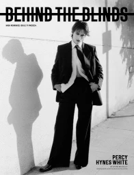 Monochrome fashion editorial portrait of a person in a tailored black suit and tie standing against an outdoor concrete wall.