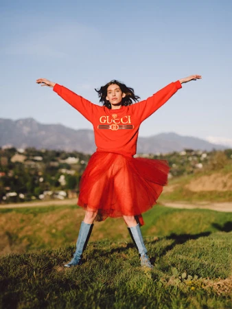 Model wearing a red Gucci sweater and red tulle skirt with blue boots standing on a grassy hill