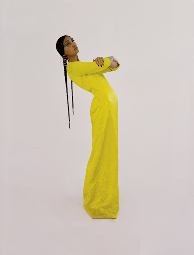 A young adult female fashion model with long braids poses with crossed arms in a fitted yellow dress against a white backdrop.