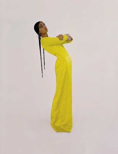 A young adult female fashion model with long braids poses with crossed arms in a fitted yellow dress against a white backdrop.