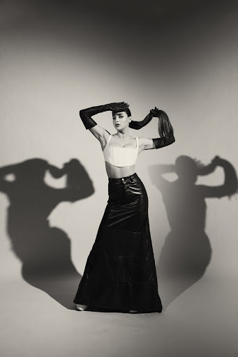 Woman wearing a white cropped top, long black leather skirt, and opera gloves against a grey studio backdrop with prominent shadows.