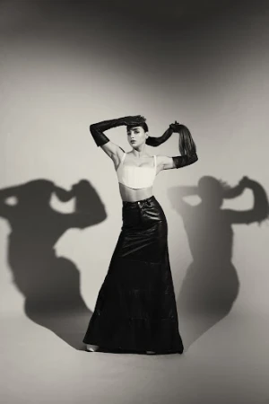 Woman wearing a white cropped top, long black leather skirt, and opera gloves against a grey studio backdrop with prominent shadows.