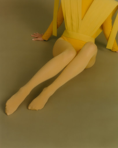 Woman in a yellow bodysuit and nude-colored tights sits on the floor in a studio with soft lighting, creating a minimalist, stylish fashion image.