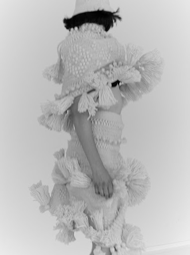 Person wearing a knitted outfit with tassels and a bucket hat, standing in front of a plain white background, fashion photography.