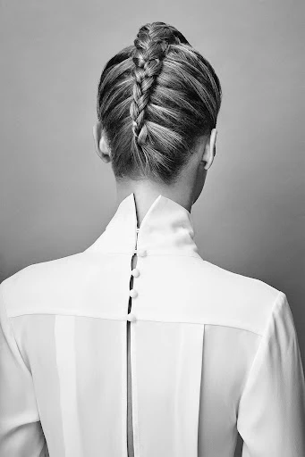 Back view of a woman with a blonde braided hairstyle wearing a white high-necked button-down blouse in a studio setting.