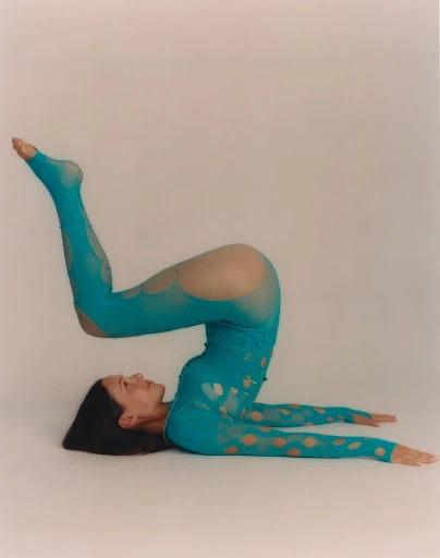 Athletic woman in a teal bodysuit performs a shoulder stand pose on a white background in a fitness or fashion context.