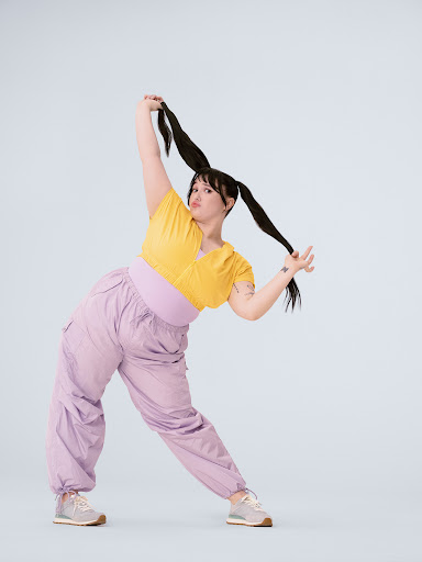 A woman in yellow top and lilac pants strikes a playful pose with pigtails against a light background in a full-length studio shot.