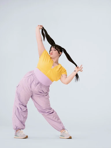 A woman in yellow top and lilac pants strikes a playful pose with pigtails against a light background in a full-length studio shot.