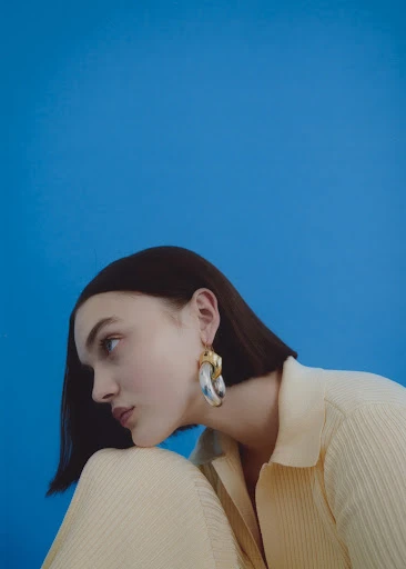 Brunette woman in a light yellow collared shirt, glancing to the side with large earrings, posing against a solid blue background.