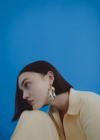 Brunette woman in a light yellow collared shirt, glancing to the side with large earrings, posing against a solid blue background.