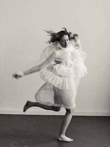 Woman in motion wearing a flowing white dress, captured in a black and white studio portrait with hair in motion against a stark background.