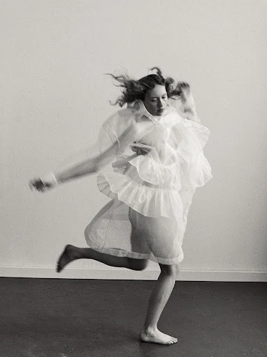 Woman in motion wearing a flowing white dress, captured in a black and white studio portrait with hair in motion against a stark background.