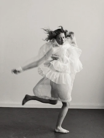 Woman in motion wearing a flowing white dress, captured in a black and white studio portrait with hair in motion against a stark background.