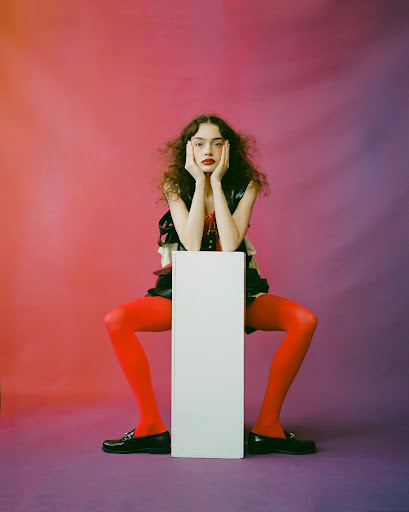 Young woman in bright red tights and dark loafers posing behind a vertical white pedestal against a vibrant gradient backdrop
