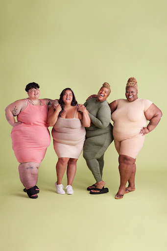 Four plus-size women stand arm-in-arm smiling against a pale green backdrop wearing casual dresses and jumpsuits in a studio setting.