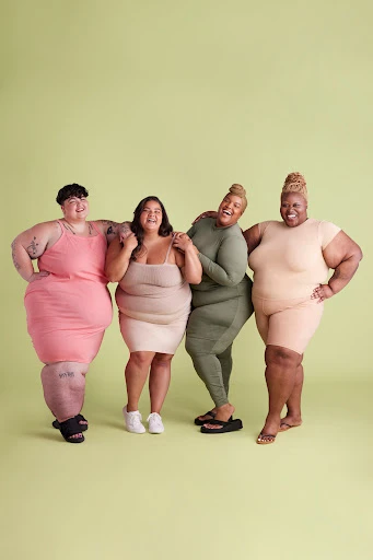 Four plus-size women stand arm-in-arm smiling against a pale green backdrop wearing casual dresses and jumpsuits in a studio setting.