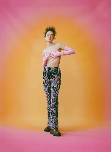 Full-length portrait of a young adult woman in a studio with patterned pants and pink gloves, striking a fashion pose on a gradient background.