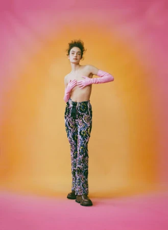 Full-length portrait of a young adult woman in a studio with patterned pants and pink gloves, striking a fashion pose on a gradient background.