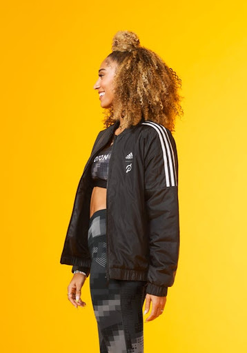 A woman with curly hair smiles while posing in athletic apparel. She's wearing an Adidas jacket and athletic top in front of a yellow background.