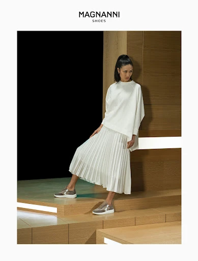 Woman in white top and pleated skirt poses with gold sneakers on lighted steps. Fashion shot for Magnanni Shoes, neutral tones.