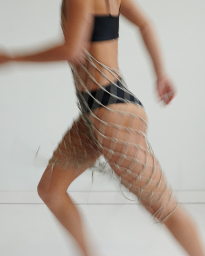 Woman wearing a black bikini and a netted garment, motion blurred against a white background. The photo captures movement and texture.