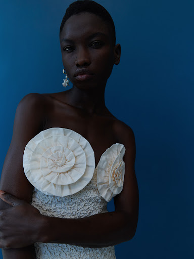 Close-up of a Black woman in a cream-colored dress with a large rosette detail, against a solid blue backdrop in studio lighting.