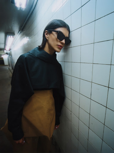 Brunette woman in sunglasses, cropped black sweatshirt, and tan miniskirt leans against a tiled wall in a dimly lit underground tunnel.
