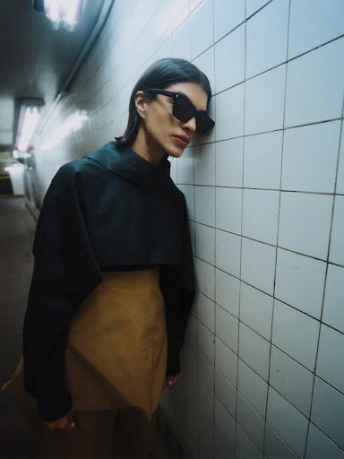 Brunette woman in sunglasses, cropped black sweatshirt, and tan miniskirt leans against a tiled wall in a dimly lit underground tunnel.
