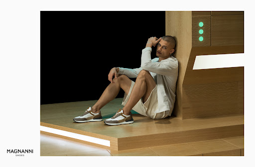 A man wearing a long-sleeved shirt and shorts sitting in front of a wooden cabinet with the Magnanni Shoes logo in the lower-left corner.