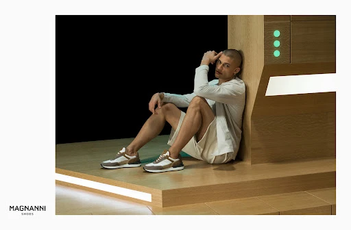 A man wearing a long-sleeved shirt and shorts sitting in front of a wooden cabinet with the Magnanni Shoes logo in the lower-left corner.