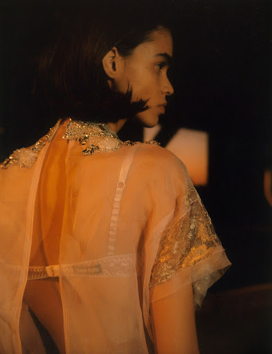 Side profile of an adult female in a peach, semi-sheer top with metallic detailing, dark short hair, and light skin against a dark background.