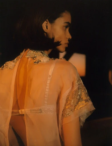Side profile of an adult female in a peach, semi-sheer top with metallic detailing, dark short hair, and light skin against a dark background.