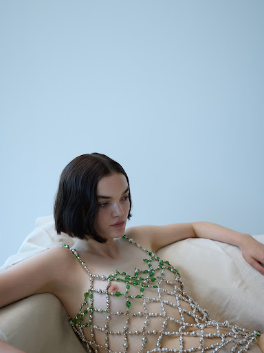 Adult female in a beaded dress is lounging on a couch in front of a light-blue wall. The portrait shot captures the model's gaze.