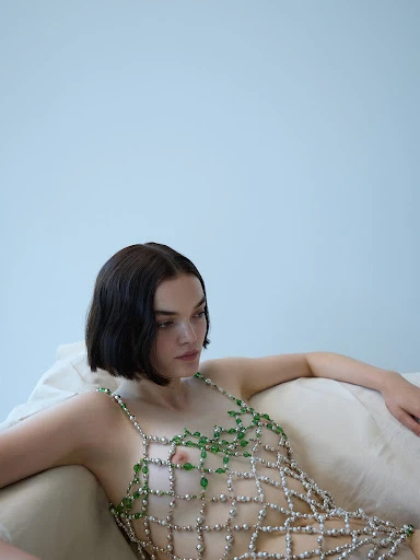 Adult female in a beaded dress is lounging on a couch in front of a light-blue wall. The portrait shot captures the model's gaze.