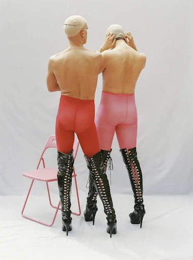 Two figures in red and pink tights and high-heeled boots stand back to back in a studio against a white backdrop. A pink chair is visible.