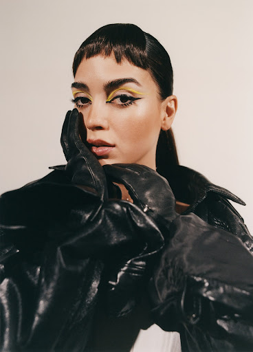 Close-up shot of a woman with yellow eye makeup, wearing a shiny black leather jacket and gloves against an off-white backdrop.