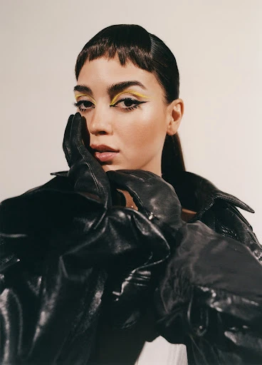 Close-up shot of a woman with yellow eye makeup, wearing a shiny black leather jacket and gloves against an off-white backdrop.