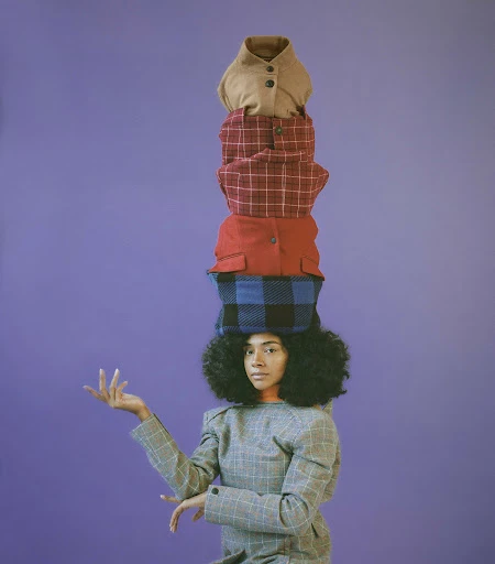 A person with an afro poses in a gray plaid dress, balancing a stack of four colorful jackets on their head against a lavender background.