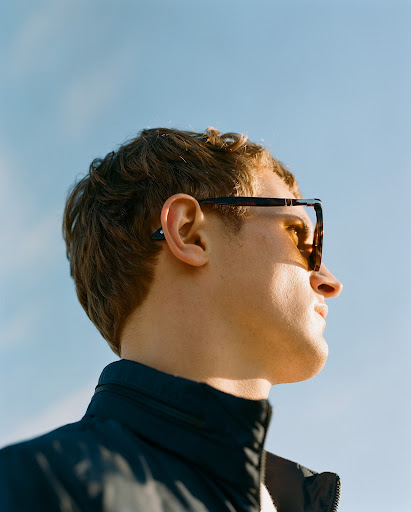 Profile of a young adult male with blond curly hair wearing sunglasses and a jacket, set against a clear blue sky.