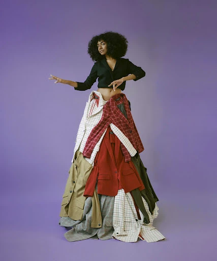 Woman poses in a fashion portrait standing against a purple backdrop, wearing a dark blouse and a skirt made of stacked coats.
