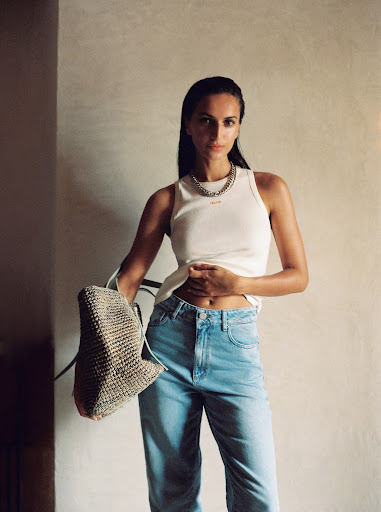 Woman wearing tank top and jeans holding a straw bag, posing in front of a textured wall. Fashion, lifestyle, studio lighting.