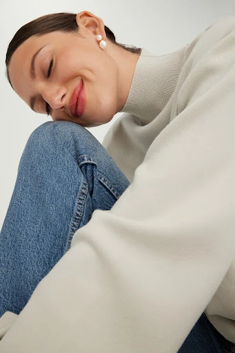 Smiling woman with closed eyes in a relaxed pose, wearing a beige sweater and jeans in a bright studio setting. Elegant pearl earrings.